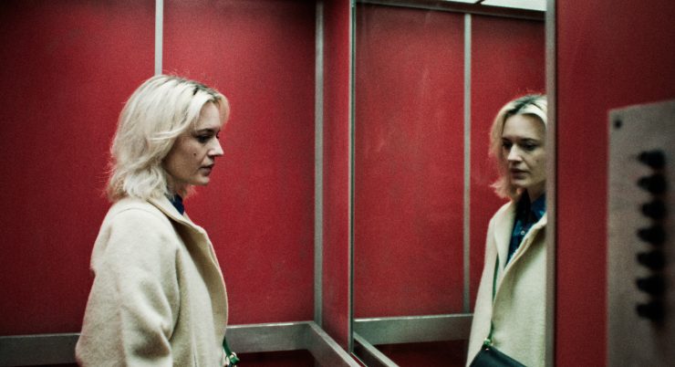 Filmstill aus 'Perla':Eine Frau mit hellblondem Haar und beigem Mantel steht in einem roten Fahrstuhl und blickt nachdenklich in den Spiegel.