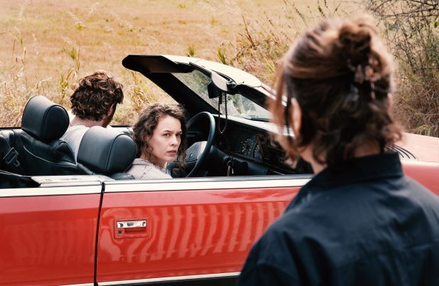 Filmstill aus Miroirs No. 3: Frau blickt aus einem roten Cabrio zu einer Person im Vordergrund - ländliche Umgebung