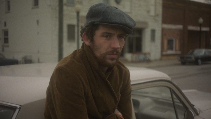 Filmstill: Ein Mann lehnt an einem Auto, sein Blick ist trüb. Er trägt eine Mütze und seine Arme sind verschränkt.