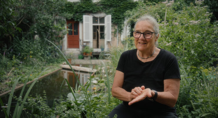 Ältere Person mit kurz geschnittenem grauem Haar und schwarzem T-Shirt sitzt in einem Garten vor einem Teich und einem Haus mit weißen Fensterläden